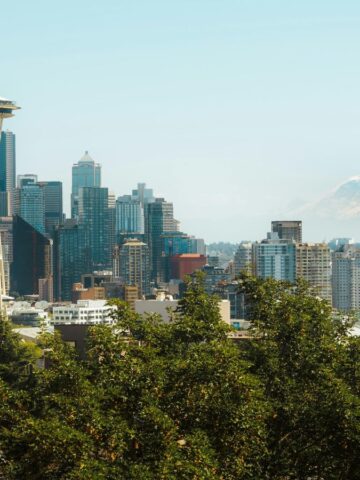 Seattle's skyline, boasting the iconic Space Needle and modern skyscrapers under a clear blue sky, is a perfect backdrop for culinary travel. With Mount Rainier in the distance and lush green trees in the foreground, it's also a hub for food tourism.