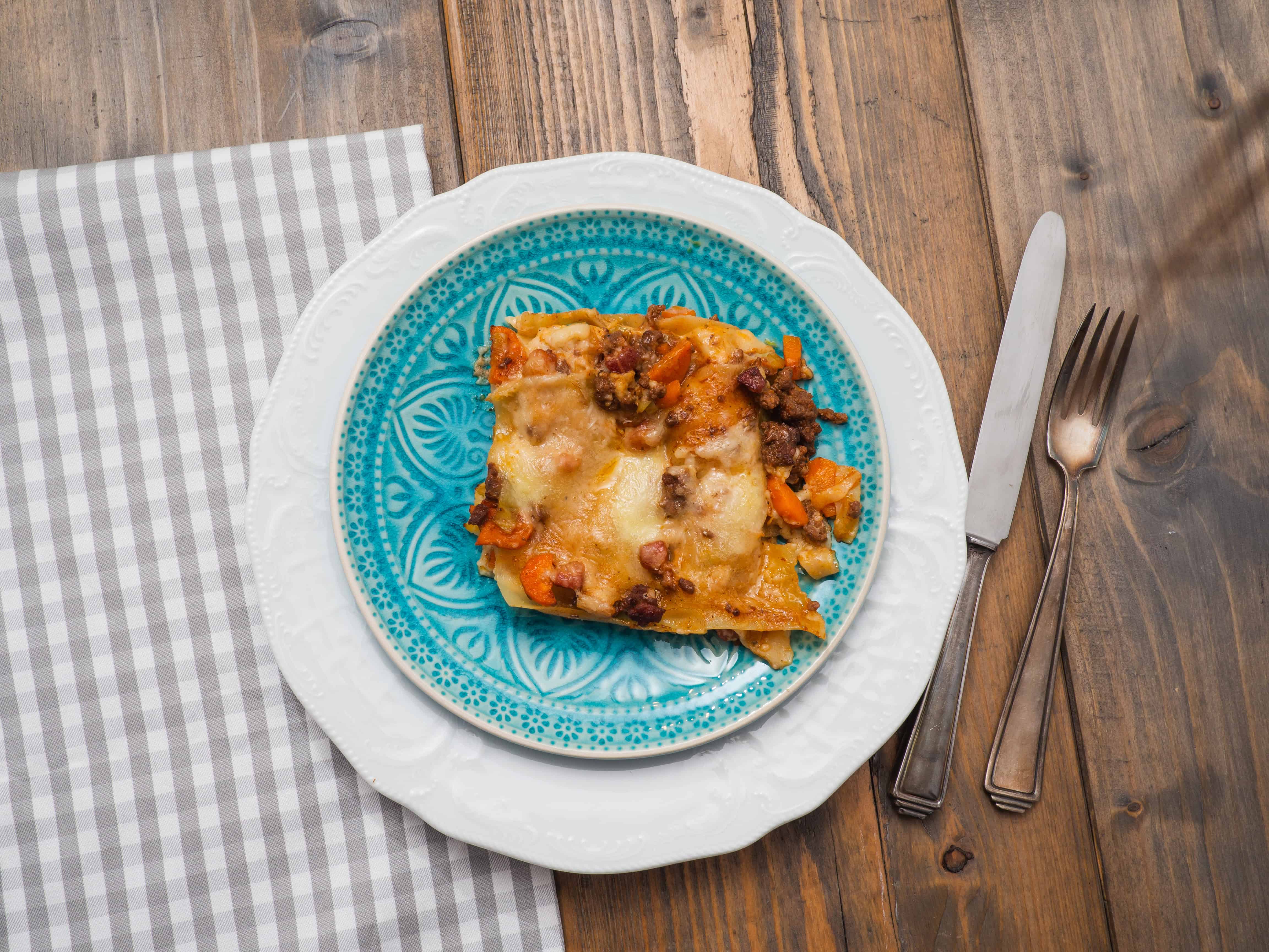 A serving of lasagna topped with cheese and ground meat sits on a blue-patterned plate, placed on a white plate, next to a fork and knife on a wooden table with a gray and white checkered napkin—an inviting twist on tropical cuisine.