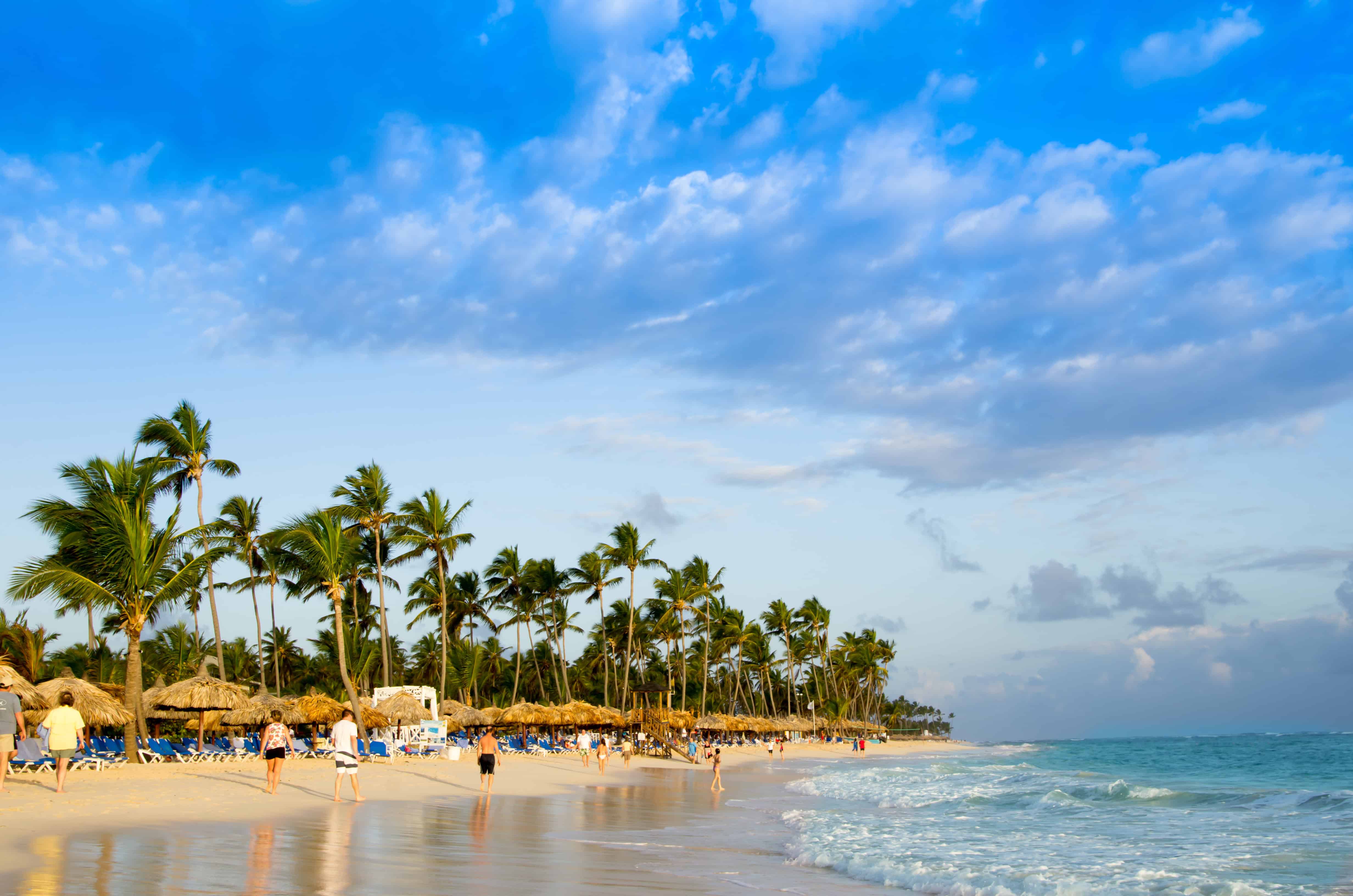 A sandy, tropical beach with people walking and relaxing, palm trees lining the shore, blue sky with scattered clouds, and gentle ocean waves.