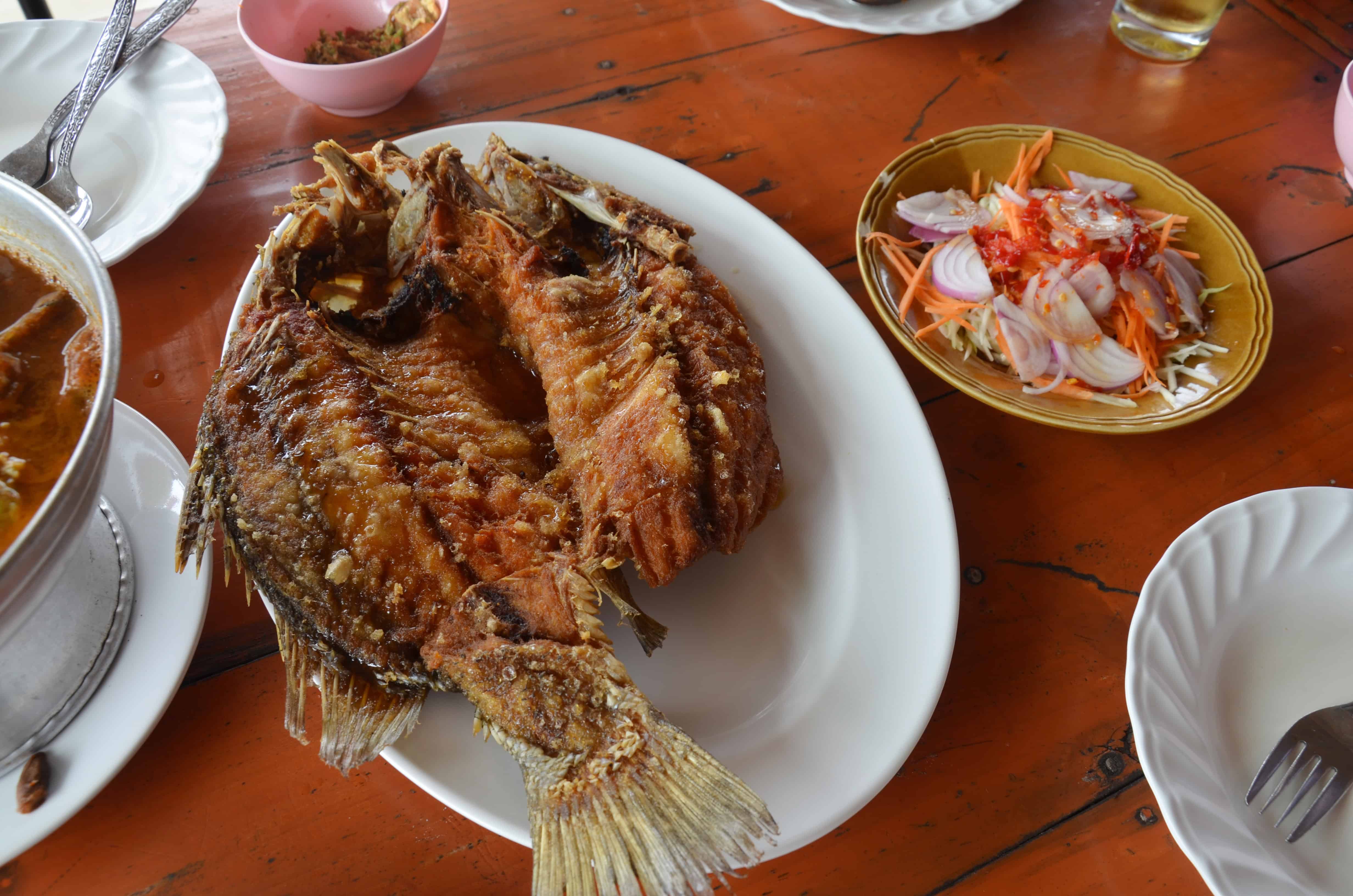 A whole fried fish, a staple of tropical cuisine and popular Dominican dishes, is served on a white plate next to sliced onions, carrots, and chili. The meal sits on a wooden table with other bowls and plates partially visible.