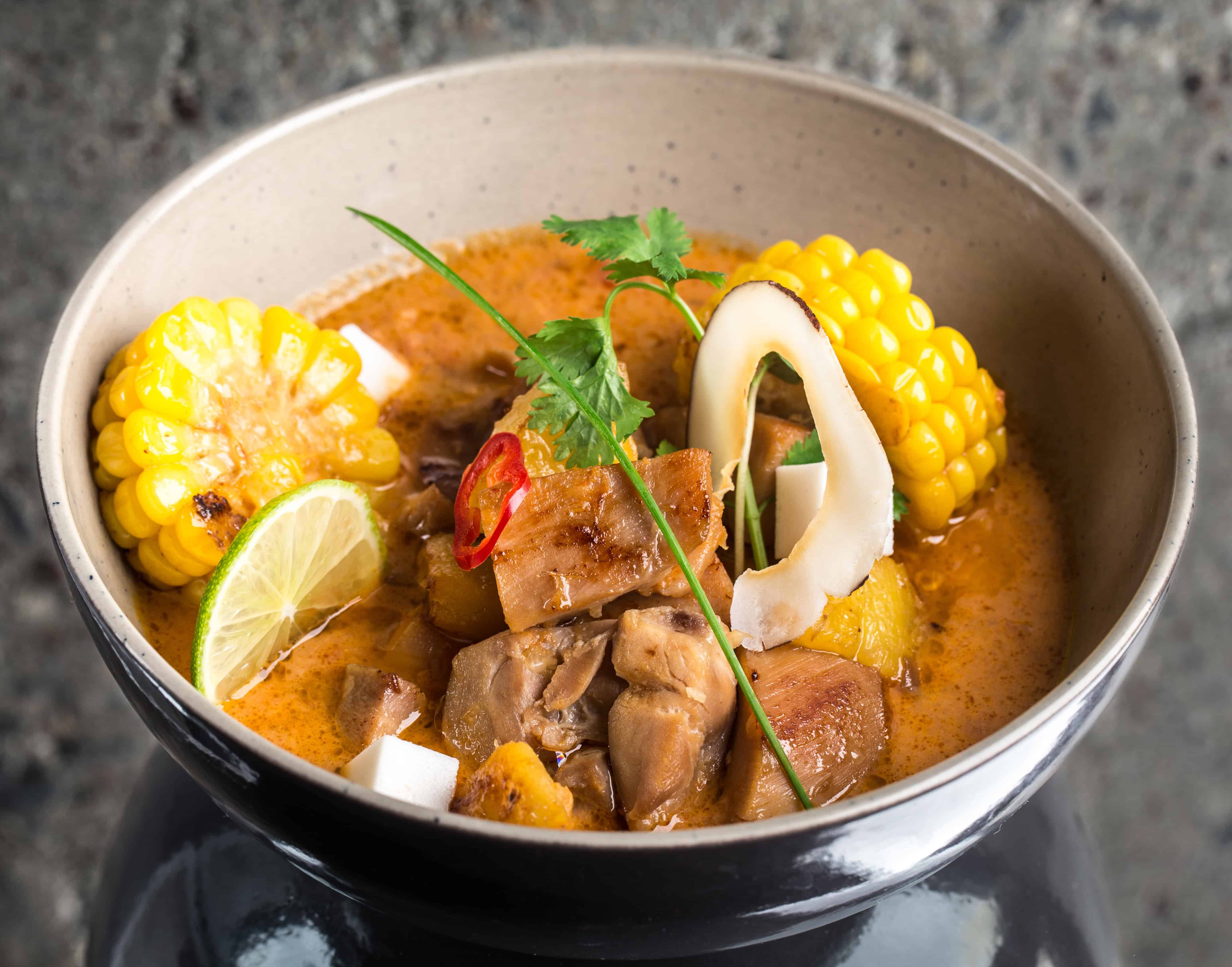 A bowl of orange-colored soup featuring chunks of meat, corn on the cob, a slice of lime, coconut pieces, and cilantro—this vibrant dish showcases tropical cuisine and is a favorite among popular Dominican dishes.