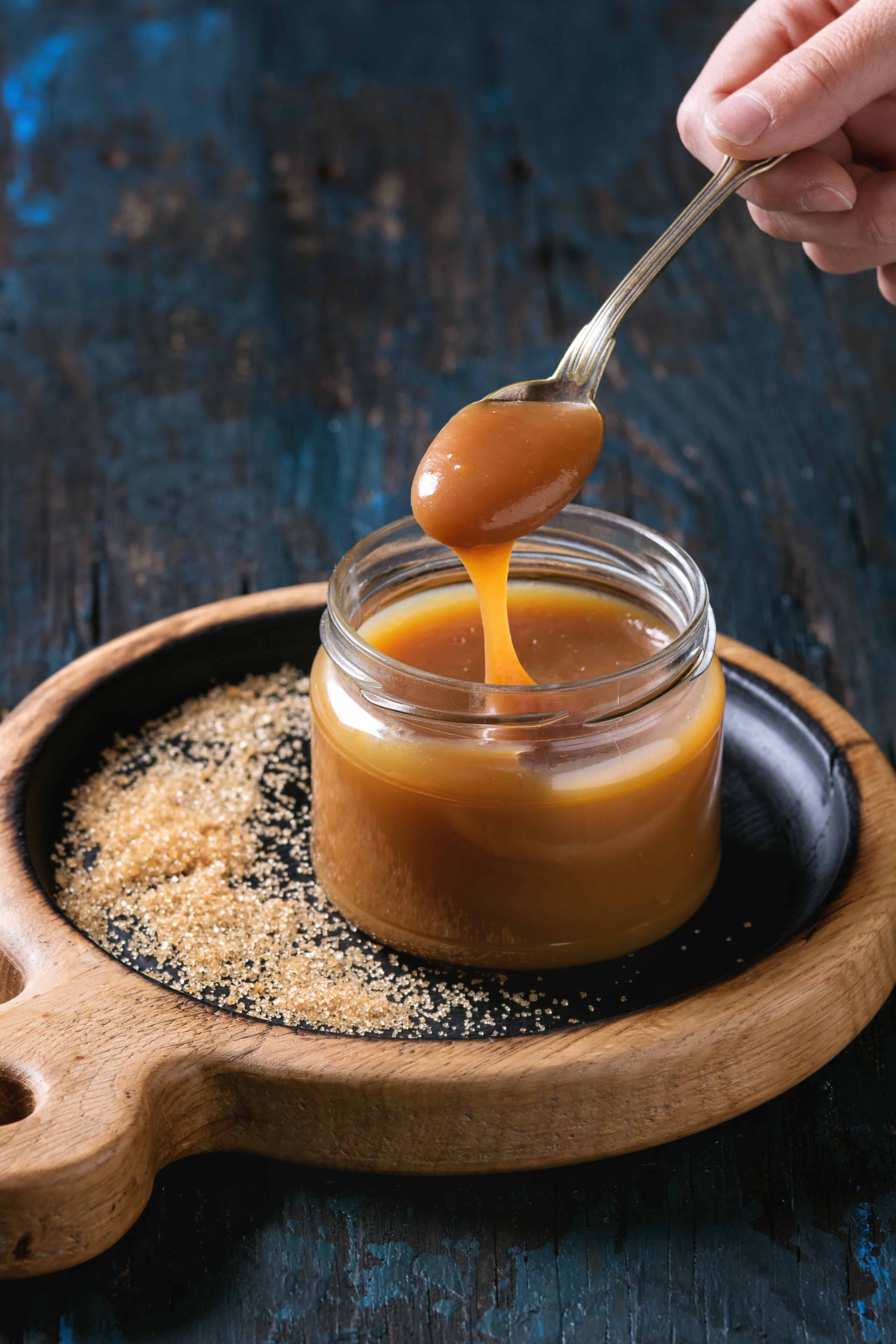 A hand lifts thick caramel sauce from a glass jar on a black plate with scattered brown sugar, set on a wooden tray against a dark background—a tempting scene inspired by the rich sweetness of Dominican Republic food.