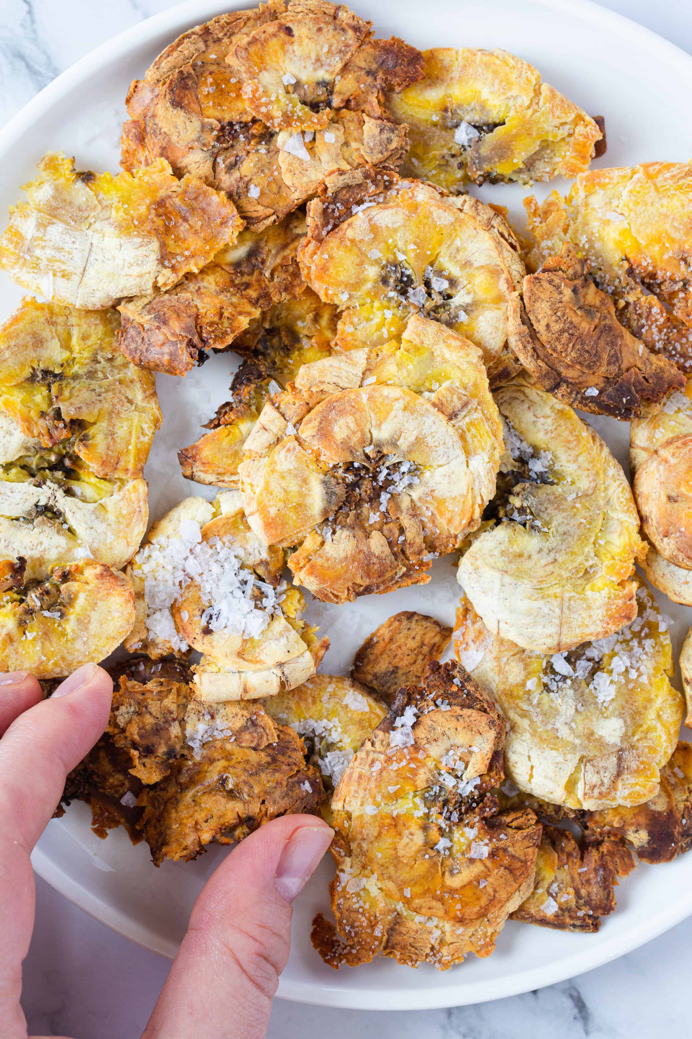 A white plate filled with round, golden-brown smashed plantain chips—a classic of Dominican Republic food—sprinkled with coarse salt. A hand is reaching in from the bottom left corner to pick up one of the chips.