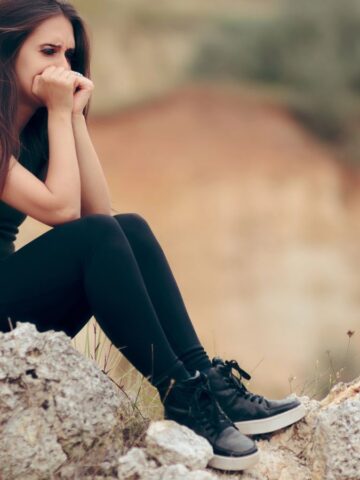 A young woman sits on rocky terrain outdoors, wearing a backpack, black leggings, and black shoes. She rests her chin on her hands and appears deep in thought. The background is blurred natural scenery.