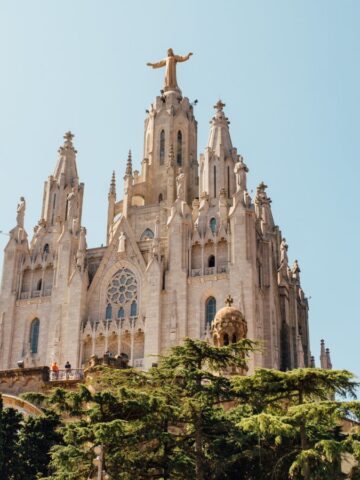 A large Gothic-style church, found in one of the Cities With the Most Unique Architecture, features spires and a tall statue of Jesus Christ on top. Its ornate facade and arched windows rise above green trees under a clear blue sky.