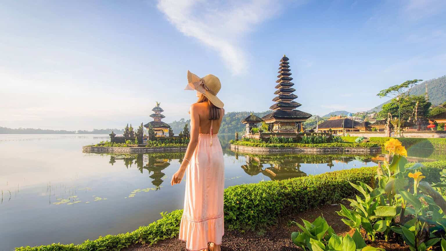 A woman in a long pink dress and wide-brimmed hat stands by a lake, looking at a traditional Balinese temple with tiered roofs, surrounded by greenery and water, under a clear sky.