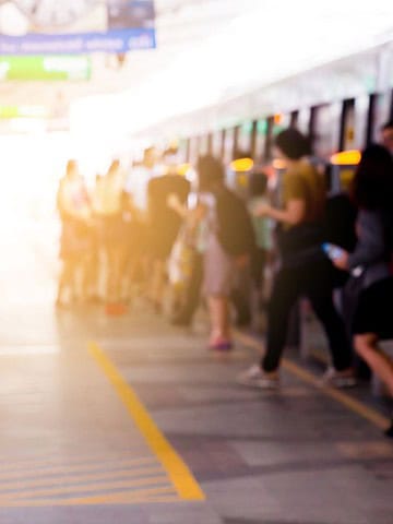 People are waiting in line and boarding a train at a station platform. The image is slightly out of focus, and sunlight is shining from the left side. Signs and markings are visible on the ground and walls.