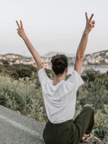 A person sits cross-legged on the side of a road, facing away from the camera, with arms raised and making peace signs with both hands. There are green bushes, hills, and a town or city in the background.
