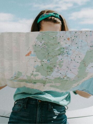 A person stands in front of a white car holding and looking at a large unfolded map. The background shows a dry, open landscape with mountains and partly cloudy sky.