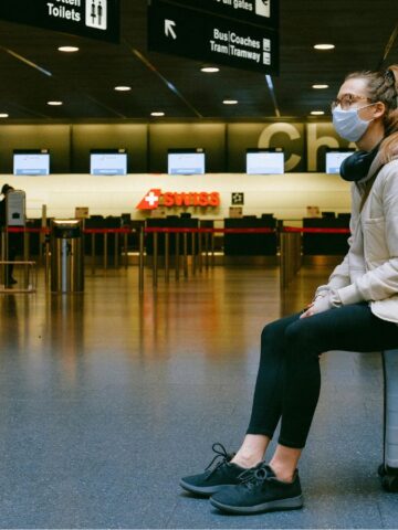 A woman wearing a face mask sits on a suitcase in an airport terminal, with check-in counters and signage visible in the background. She has headphones around her neck and a backpack on her back.