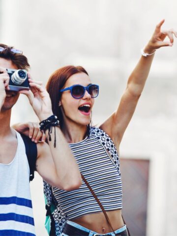 A young man in a striped tank top takes a photo with a camera while a young woman in sunglasses and a striped crop top stands beside him smiling and pointing excitedly at something in the distance.