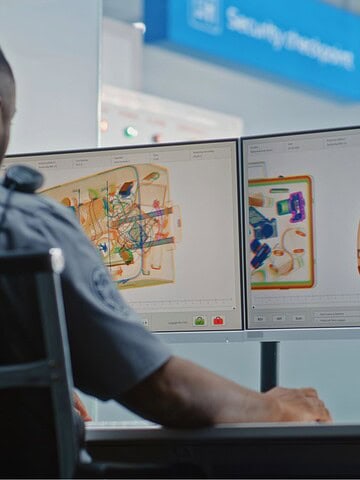 A security officer sits at a desk, monitoring two computer screens displaying x-ray images of bags at an airport security checkpoint. A person stands in the background near the screening area.