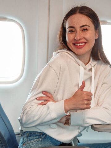 A woman with long brown hair sits by the window on an airplane, wearing a white hoodie and jeans. She smiles and gives a thumbs up, with empty seats beside her and sunlight coming through the window.