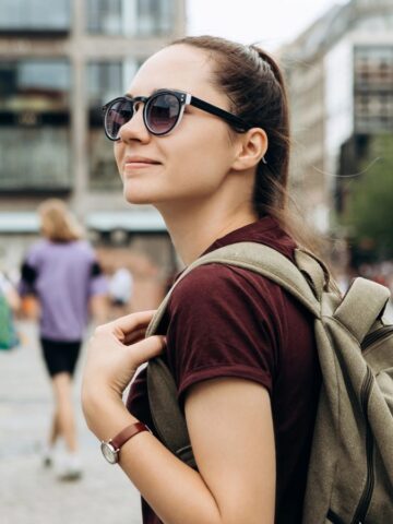A young woman wearing sunglasses and a backpack stands outdoors in a city square. Several people walk in the background. She is looking slightly upward and smiling. Buildings and trees are visible behind her.