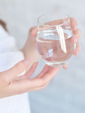 A person wearing a white towel holds a clear glass of water with both hands against a light, blurred background.