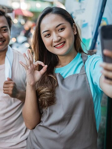 A woman wearing an apron takes a selfie and makes an OK gesture while a man stands beside her, both smiling, next to a food stall with baskets of vegetables and kitchen supplies.