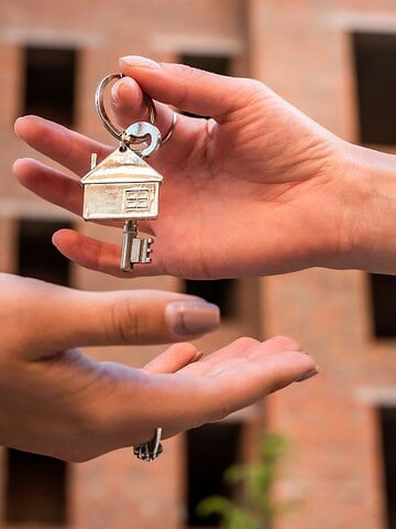 A person hands over a key with a house-shaped keychain to another person. The background shows a building under construction with visible brick walls and rectangular openings.