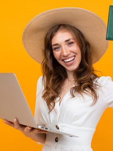 A woman wearing a wide-brimmed hat and white dress holds a laptop in one hand and raises a passport with boarding pass in the other, smiling in front of a bright orange background.