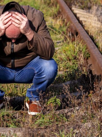 A person in a brown jacket and blue jeans squats between two rusty railway tracks, holding their head in their hands. Grass and weeds grow around the tracks.