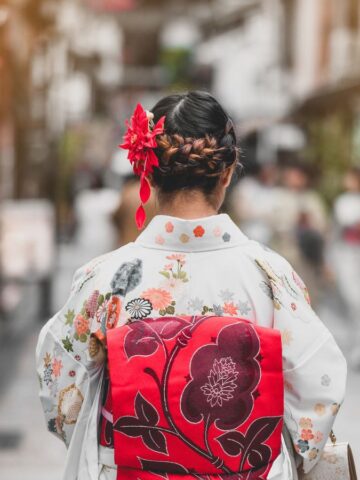 A person wearing a floral-patterned kimono with a red and pink obi stands on a street, facing away from the camera. Their hair is styled in braids with red hair accessories. The background shows a blurred street scene.
