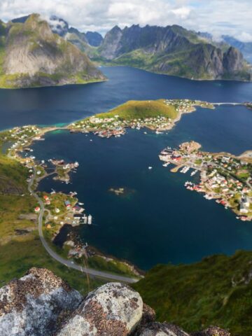 Aerial view of a coastal village surrounded by mountains and water, with clusters of houses, a harbor, winding roads, and green hills under a partly cloudy sky.