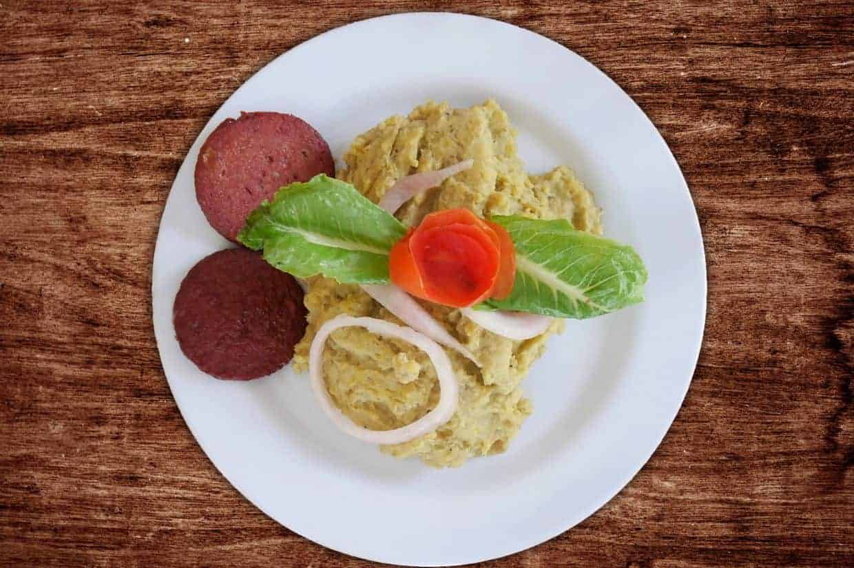 A white plate with mashed plantains, two slices of fried sausage, sliced onions, lettuce, and a tomato rose garnish—an inviting showcase of popular Dominican dishes—set on a wooden surface.