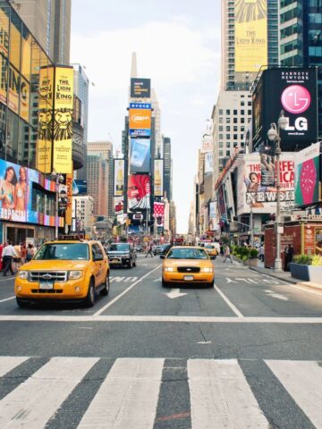 Yellow taxis drive through a busy street in Times Square, New York City, surrounded by tall buildings with colorful billboards and advertisements; crowds of pedestrians walk on both sides.