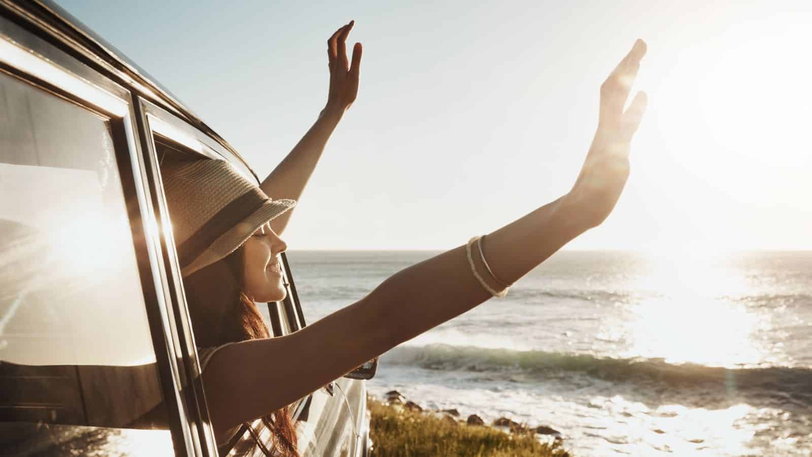 A person wearing a hat leans out of a vehicle window with both arms raised, facing the ocean and the setting sun. The scene is outdoors near a shoreline with waves and sunlight visible.