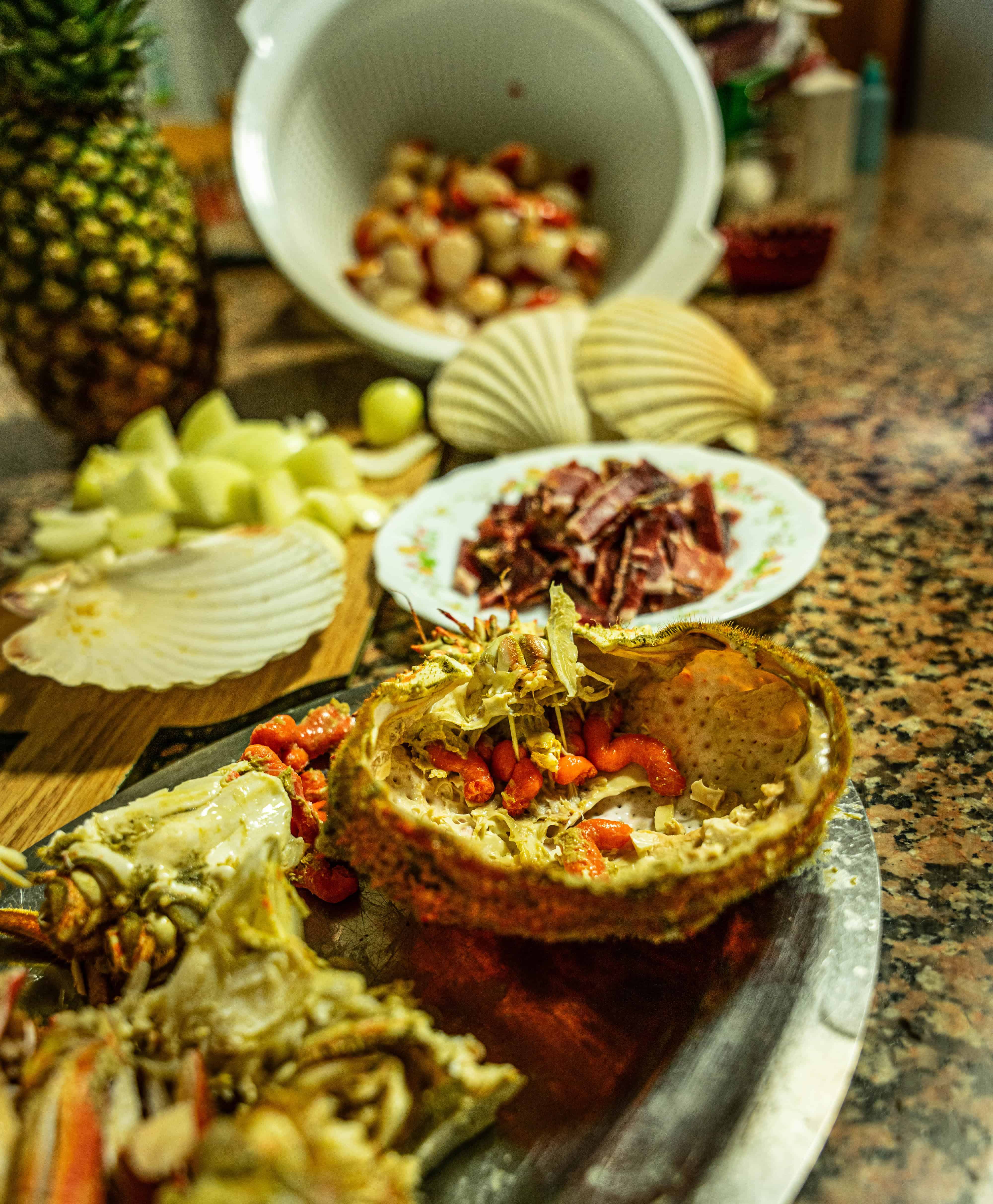 A prepared crab shell filled with crab meat sits on a metal tray, surrounded by sliced melon, shellfish, and pineapple—vibrant touches of tropical cuisine often seen in popular Dominican dishes—while a white colander rests in the background.