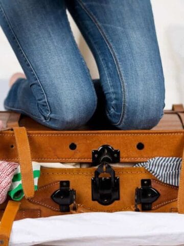 A person kneels on top of an overfilled brown suitcase, trying to close it, with clothes spilling out from the sides. The suitcase is on a white surface.