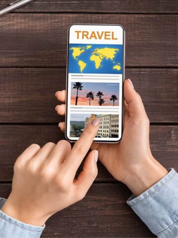 A person holds a smartphone displaying a travel app with photos of destinations. Their finger is touching the screen. A cup of coffee, a payment card, a keyboard, and a pen are on the wooden desk.