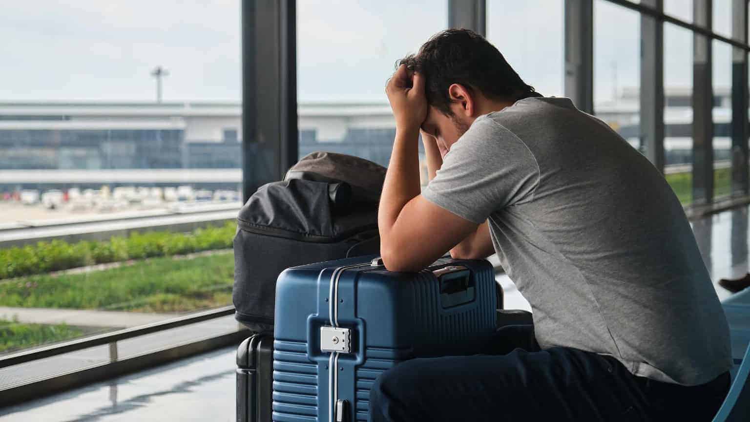 A man sits at an airport with his head in his hands, appearing stressed or upset. He is next to a blue suitcase and a backpack, looking out the large window at the runway outside.