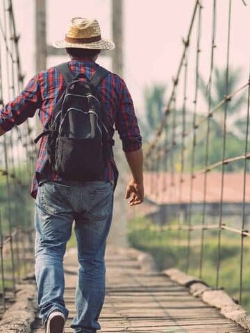 A person wearing a straw hat, plaid shirt, jeans, and a black backpack walks across a narrow suspension bridge made of wood and rope in an outdoor, rural setting with trees and buildings in the background.
