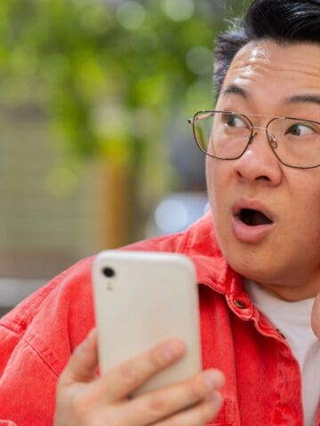 A person wearing glasses and a bright red jacket holds a smartphone in one hand, looking surprised with their mouth open and the other hand touching their cheek. The background is outdoors and slightly blurred.