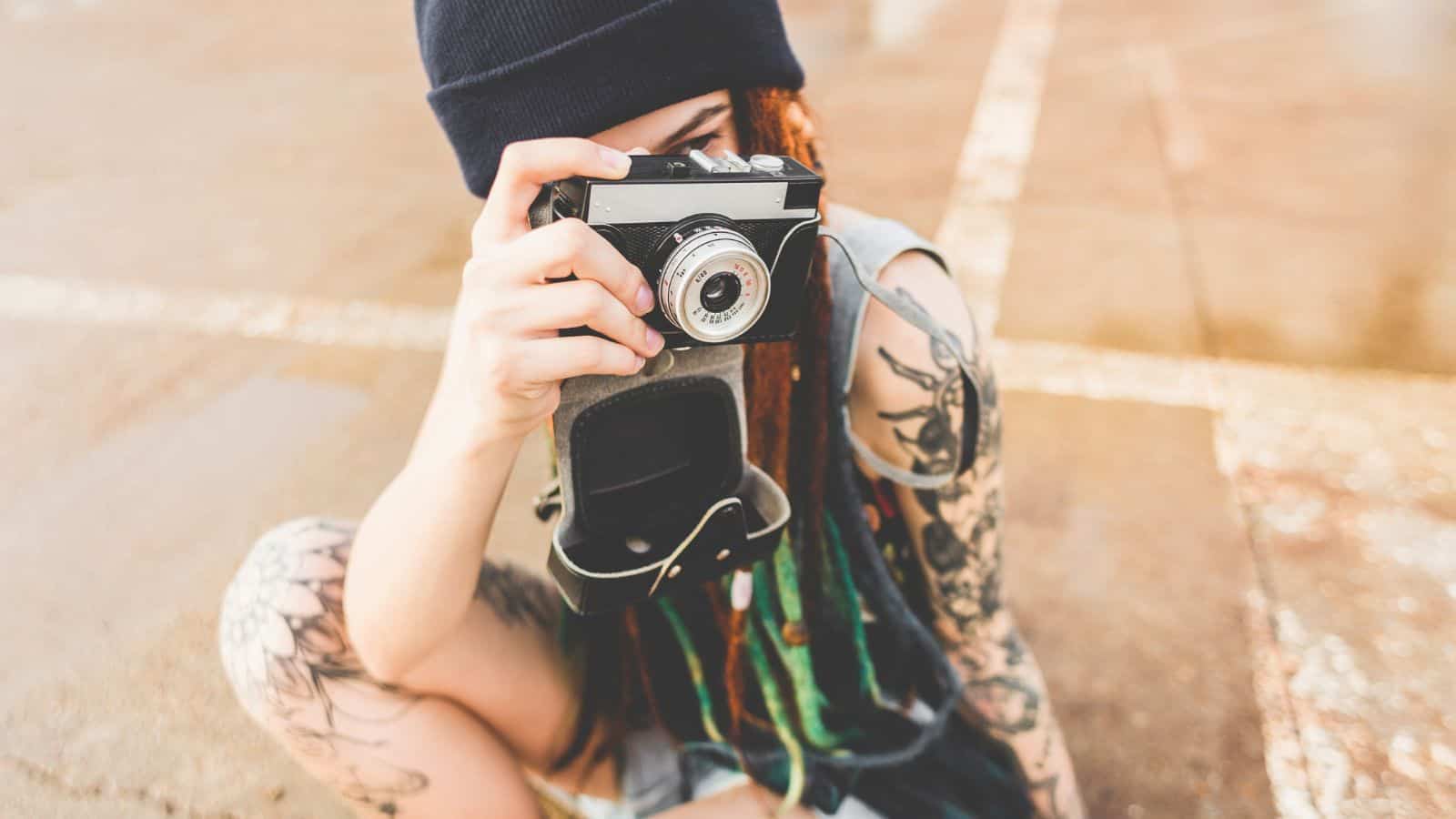 A person with tattoos and a black beanie is sitting on the ground, holding a vintage camera up to their face as if taking a photo. The camera case is open and hanging from their hand.