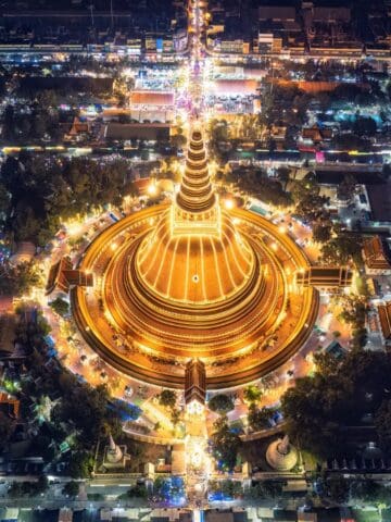 Aerial view of a large, illuminated golden pagoda at night, surrounded by trees, buildings, and busy streets with colorful lights and traffic in an urban area.