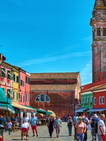 A busy street scene in a European town with colorful buildings, market stalls covered by green awnings, many people walking, and a tall, leaning bell tower in the background under a clear blue sky.