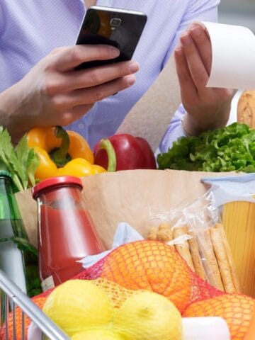 A person holding a smartphone and a receipt stands behind a grocery cart filled with vegetables, fruits, drinks, and other groceries in a supermarket.