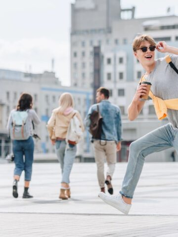 A young person wearing sunglasses and a backpack smiles while holding a coffee cup and talking on the phone. Four people with backpacks walk away in the background in an urban setting.