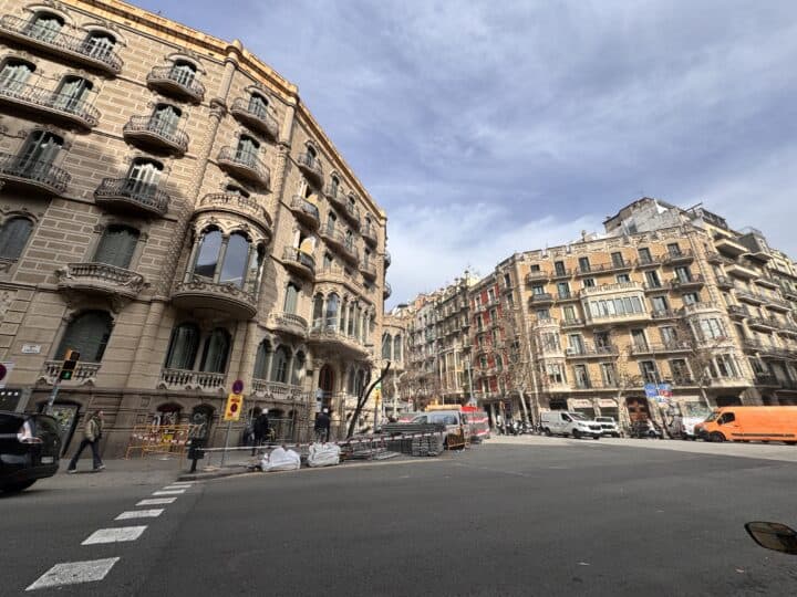 A city street intersection in Family-Friendly Barcelona, with ornate historic buildings, cars parked along the sides, construction materials near the corner, and a cloudy sky overhead.