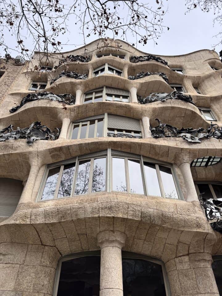 A stone building with curved, wavy lines and wrought iron balcony railings viewed from below—a great spot to admire on your Family Trip Barcelona. Leafless tree branches extend overhead against a cloudy sky.