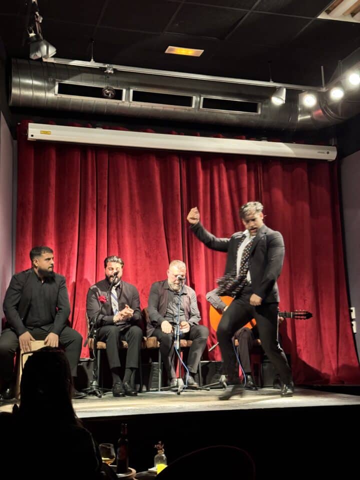 A man energetically dances flamenco on stage in front of four seated musicians dressed in black suits, set against a backdrop of red curtains—an authentic, family-friendly Barcelona experience for those enjoying Barcelona with kids.