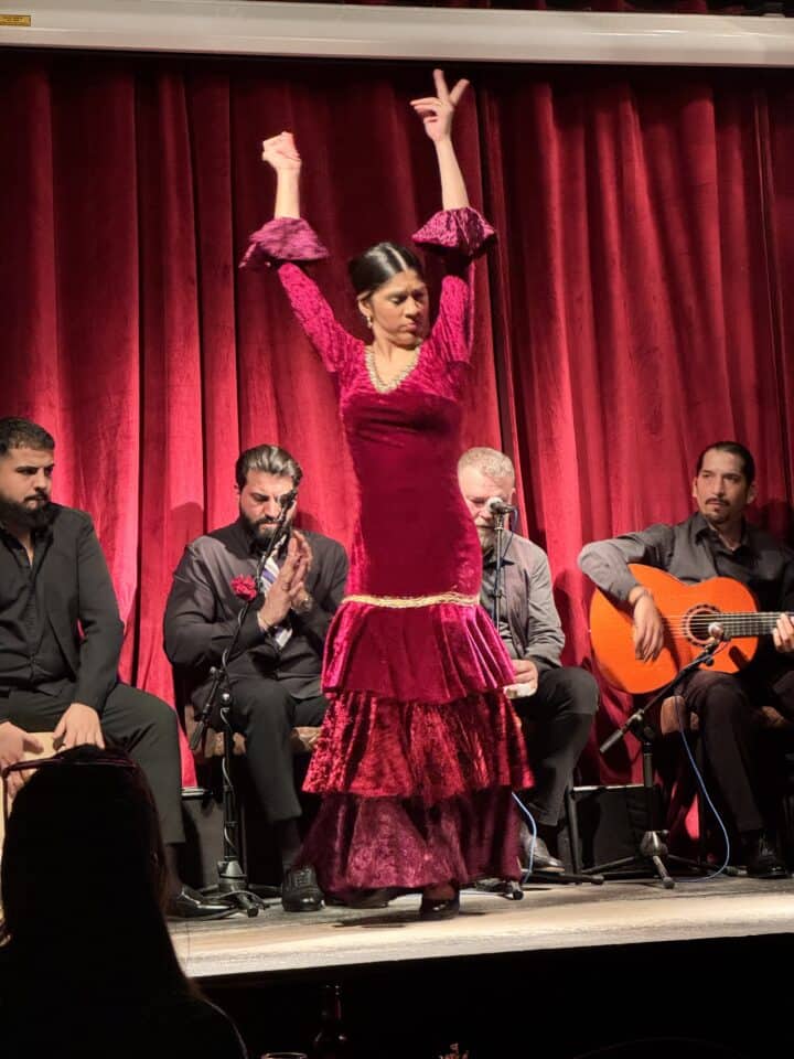 A woman in a red flamenco dress dances on stage with her arms raised, as four men sit behind her—one playing guitar. Enjoy this family-friendly Barcelona performance, set against deep red curtains, during your week in Barcelona.