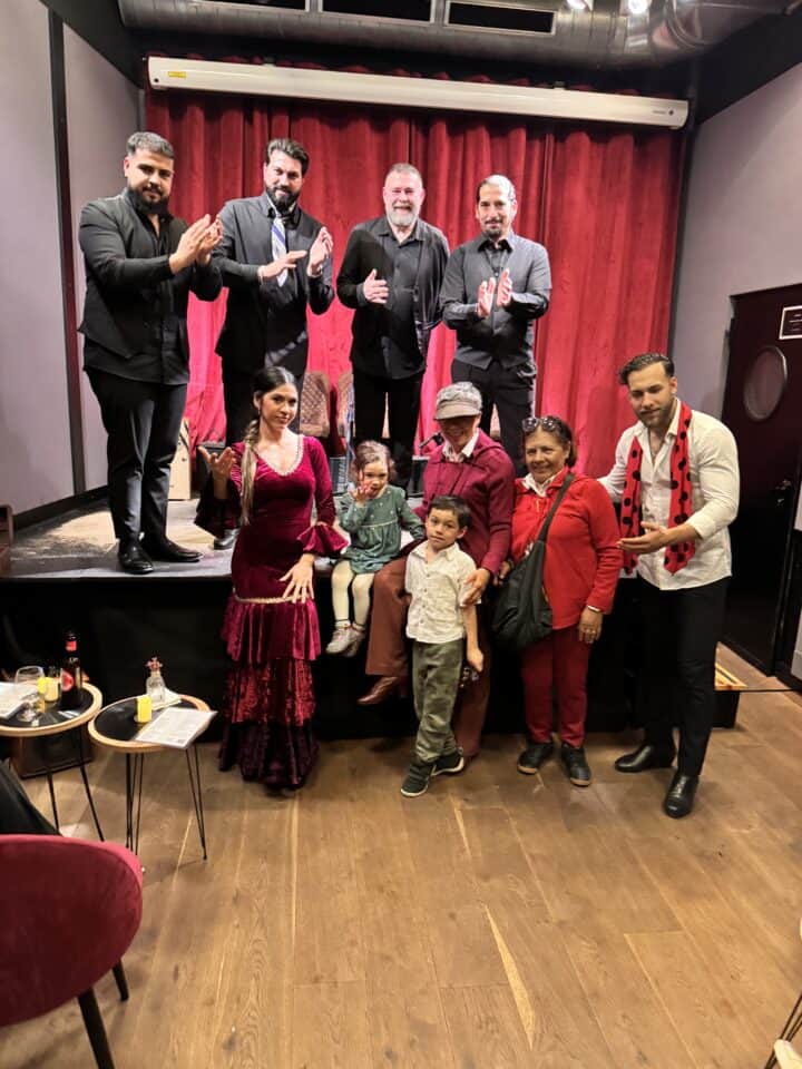 A group of eight adults and two children pose together in front of a red curtain on a stage. Perfect for a Barcelona family vacation, the scene captures laughter as some clap on stage while others, with the kids, stand by tables with drinks.
