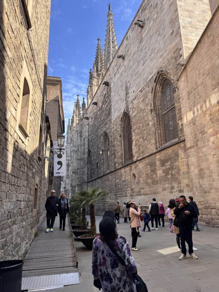 People stroll near the side of a large Gothic-style cathedral with pointed arches and spires under a blue sky. Some are taking photos, capturing memories from their Barcelona family vacation, while a banner hangs from a nearby building on the left.