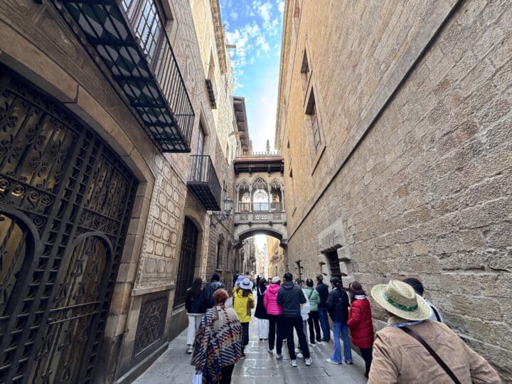 A group of people stands and walks along a narrow stone street between tall old buildings in family-friendly Barcelona, facing a decorative stone bridge connecting the structures above. The sky is partly cloudy.