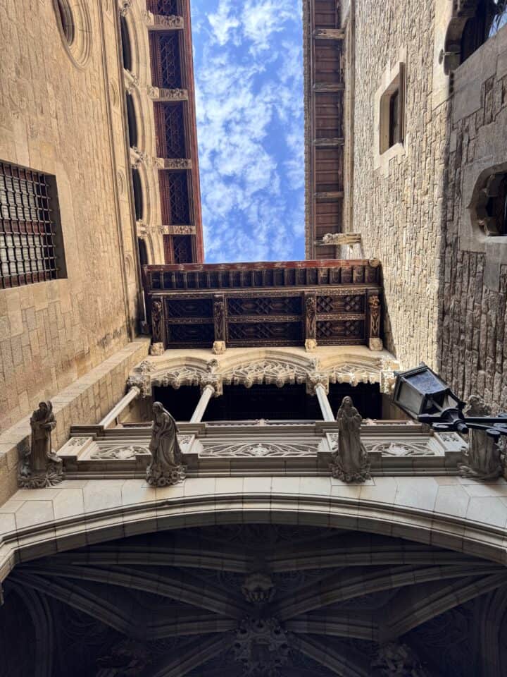 Looking up at an ornate stone bridge connecting two historic buildings in Family-Friendly Barcelona, detailed carvings and balconies stand out against beige walls and a blue sky—perfect for exploring on your Barcelona family vacation.