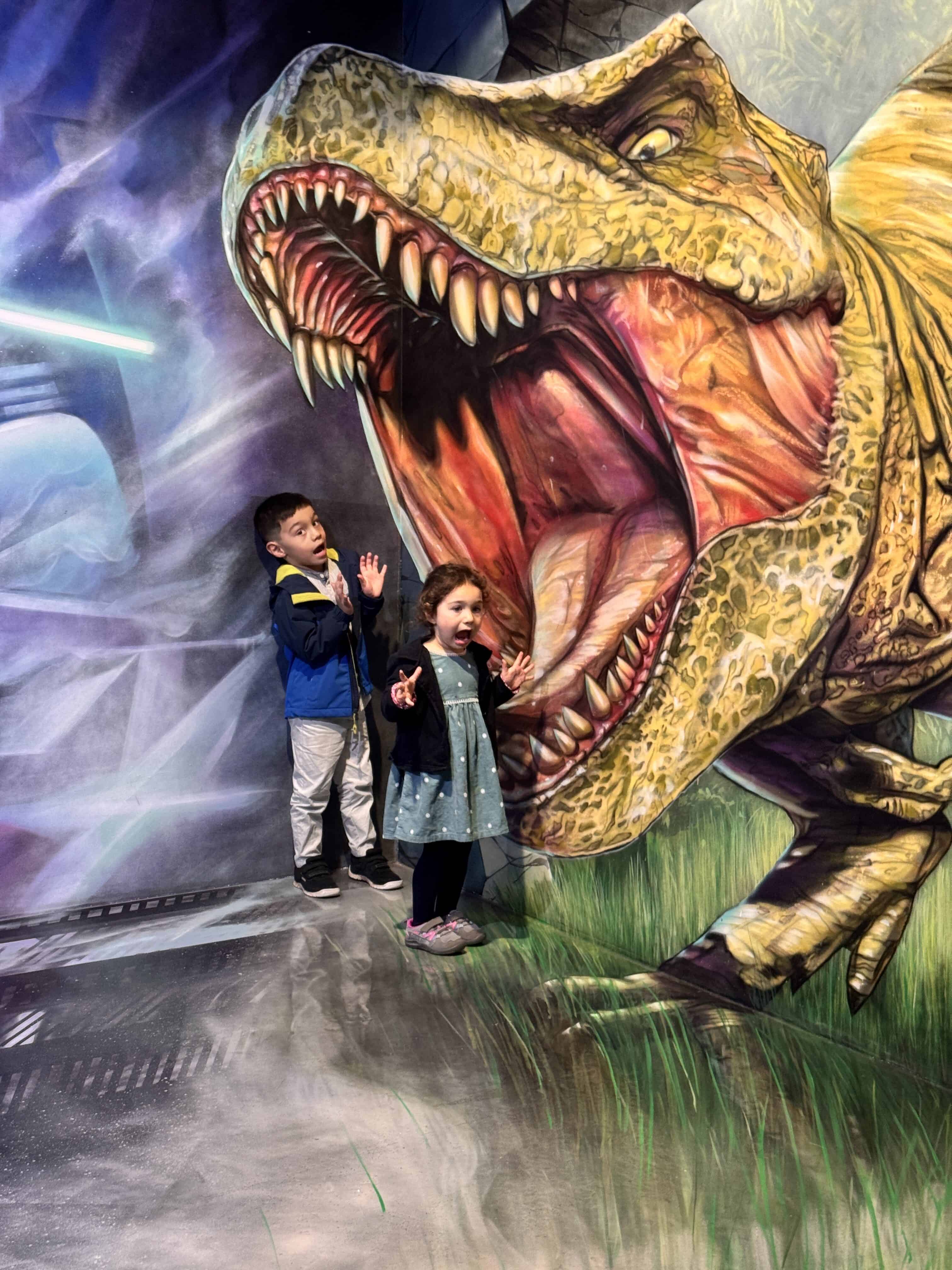 Two children pose playfully in front of a mural of a large roaring T. rex, reacting with surprise as if the dinosaur is about to eat them—a fun moment from a Family-Friendly Barcelona adventure.
