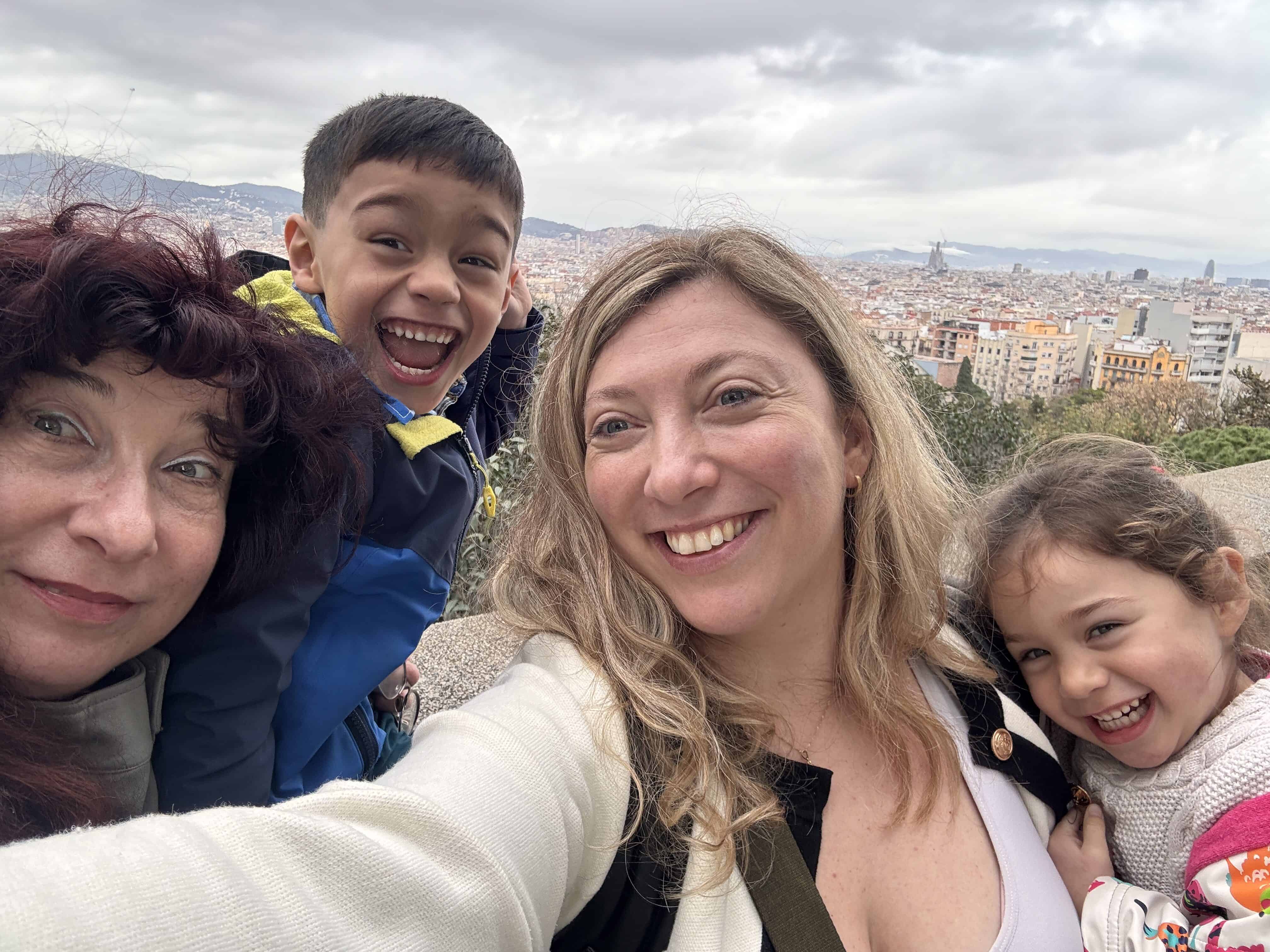Four people take a selfie outdoors with a cityscape in the background. The adults and children are smiling at the camera, capturing a joyful moment during their Barcelona family vacation under cloudy skies.