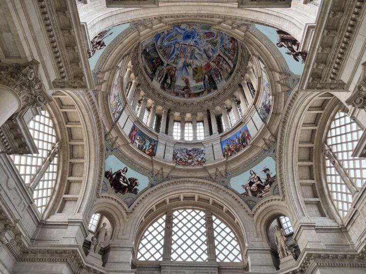 View looking up at the interior dome of a grand building in family-friendly Barcelona, with ornate architectural details, large arched windows, and colorful murals depicting various scenes and figures on the dome and surrounding surfaces.